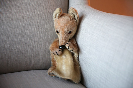 In der Ecke eines Sofas sitzt als Handpuppe ein Fuchs, der durch seine gesenkte Kopfhaltung schüchtern wirkt.