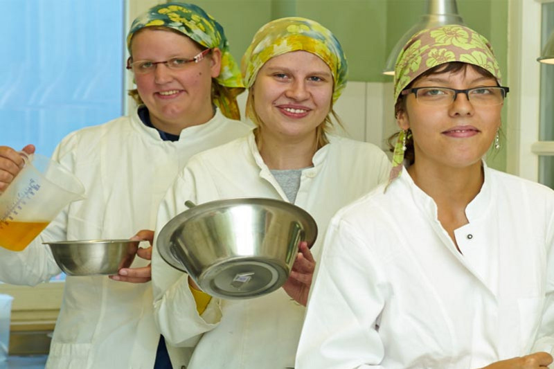 Drei junge Frauen in weißen Kitteln und Kopftüchern stehen in einer Küche