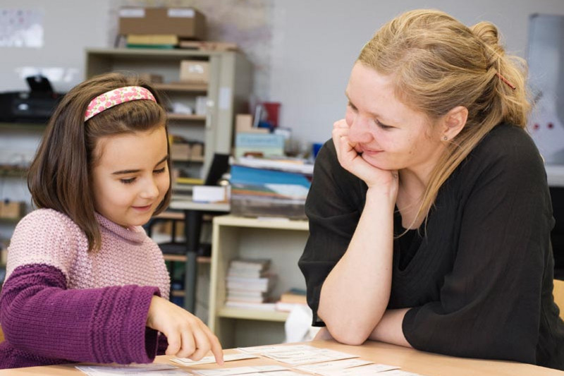 A young girl and a female teacher sitting together at a table in a class room, learning