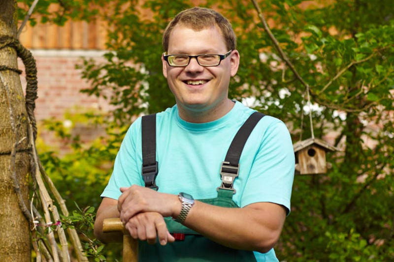 A young man in work clothes stands with a spade in a garden and smiles into the camera