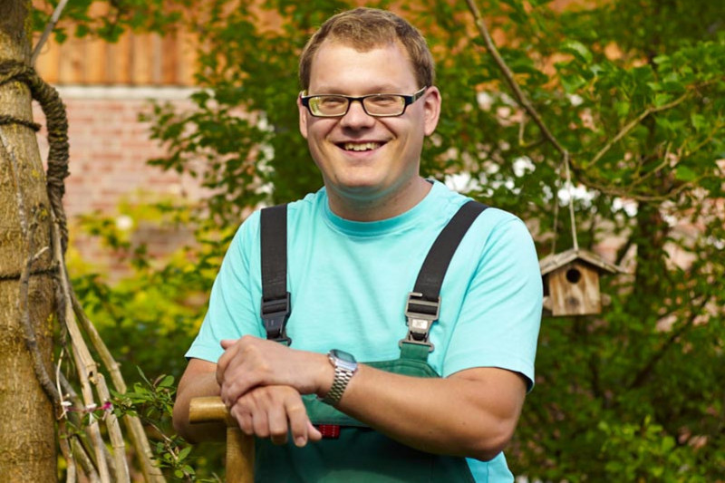 Ein junger Mann in Arbeitshose steht mit einem Spaten im Garten und lacht in die Kamera