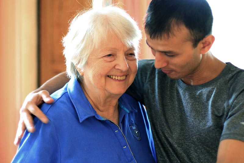 A care taker puts his arm around a older woman smiling