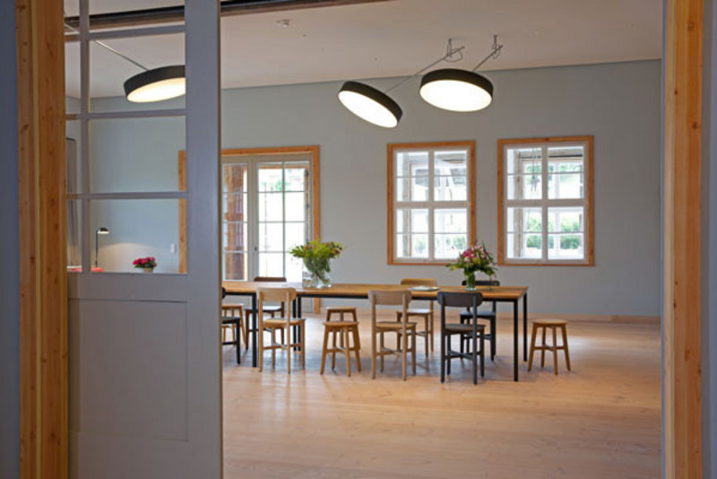 A bright room with big windows and a wooden table in the middle surrounded by chairs 